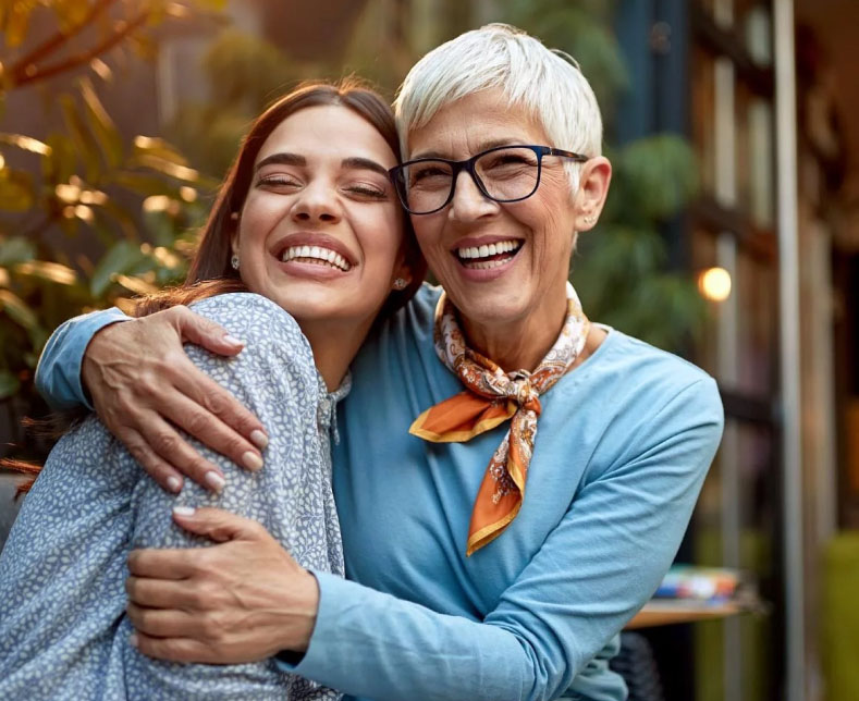 Two women smiling together in festive holiday setting with warm background lights.