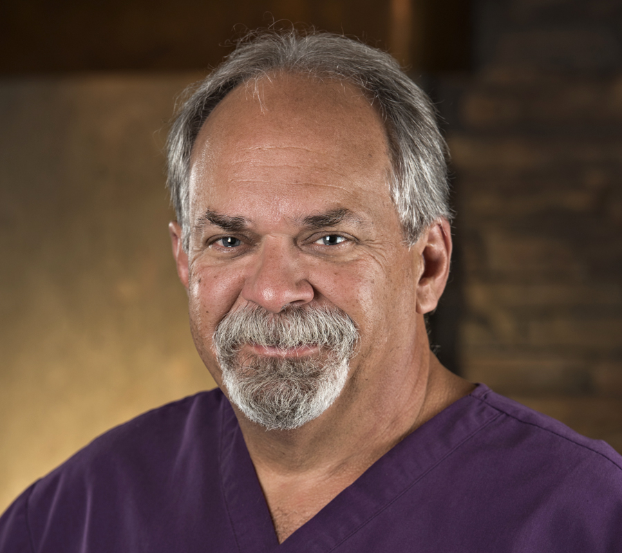 Middle-aged man with gray hair and beard wearing purple scrubs in professional medical office setting.