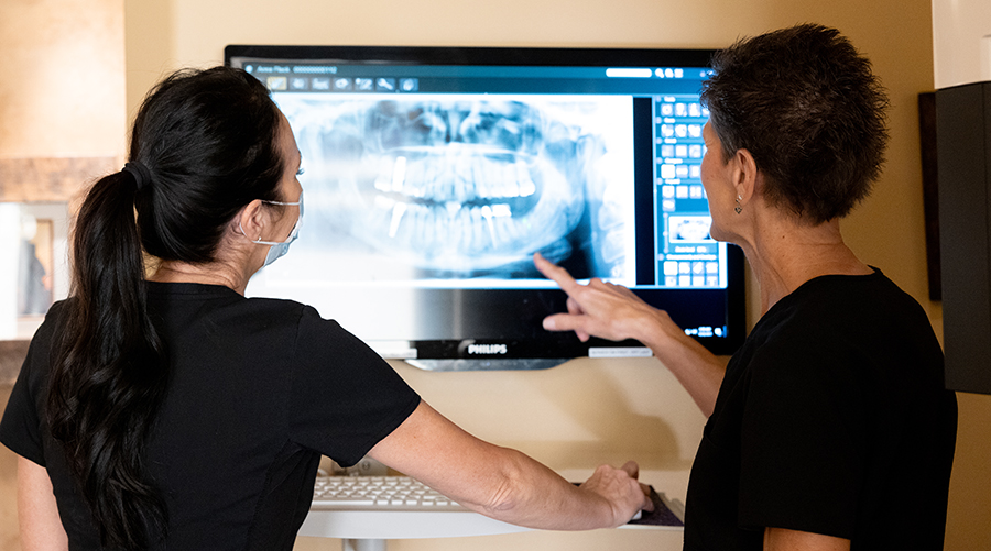 Two dental professionals examine X-ray images on a monitor in a clinical setting.