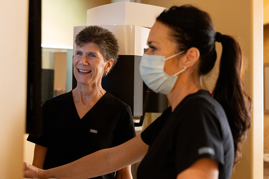 Two dental professionals in black scrubs converse in a clinical treatment room setting.