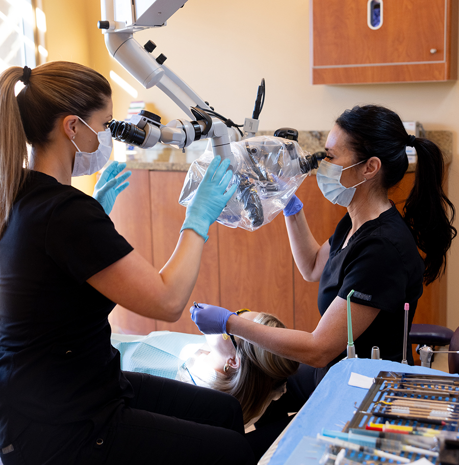 Two dental professionals wearing masks and gloves examine dental equipment in a clinical treatment room.