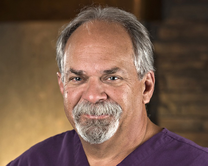 Professional headshot of male dentist in purple scrubs against warm-toned office background.